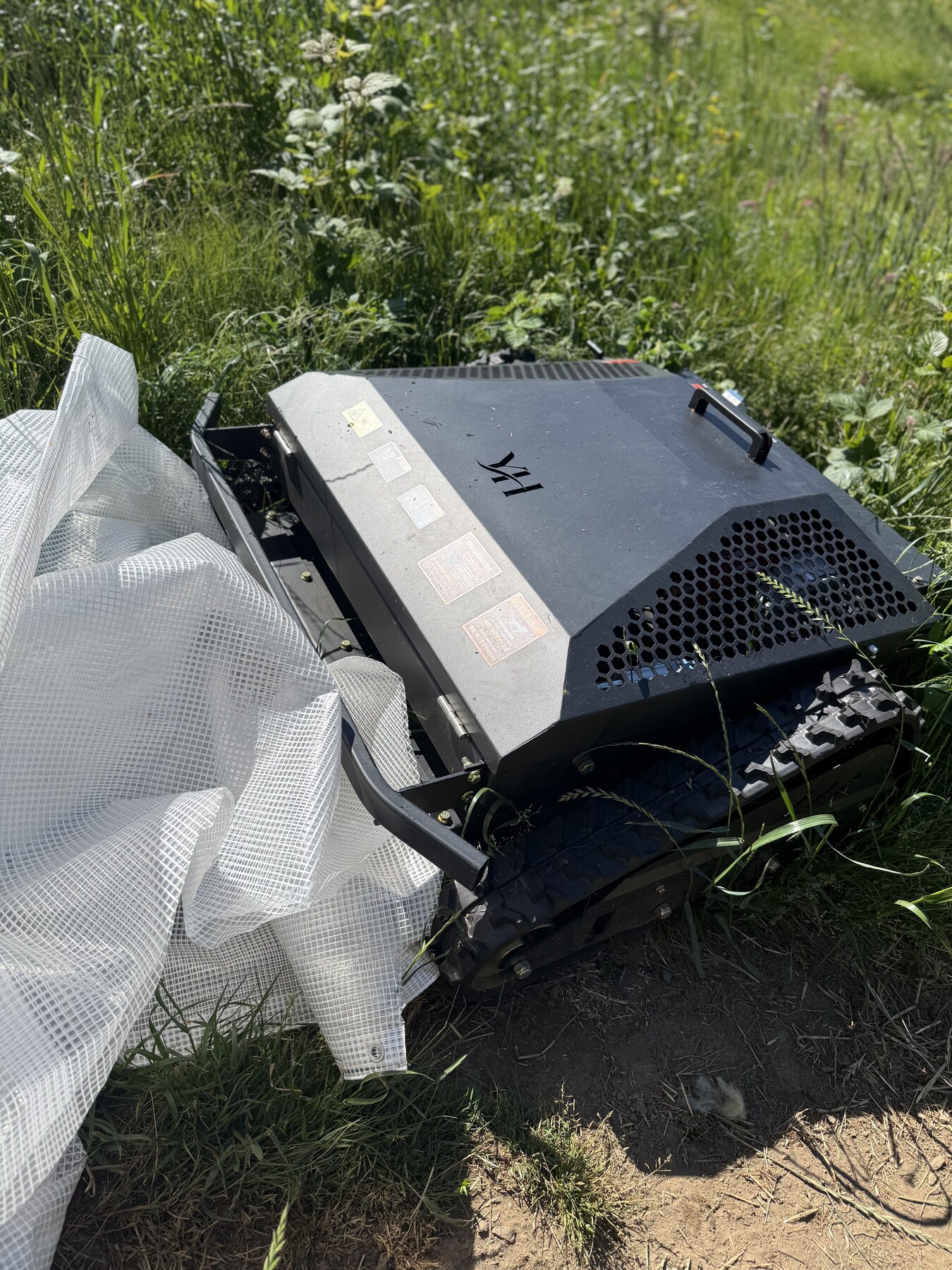 The tracked hillside mower sitting in tall grass with its protective tarp partly removed.